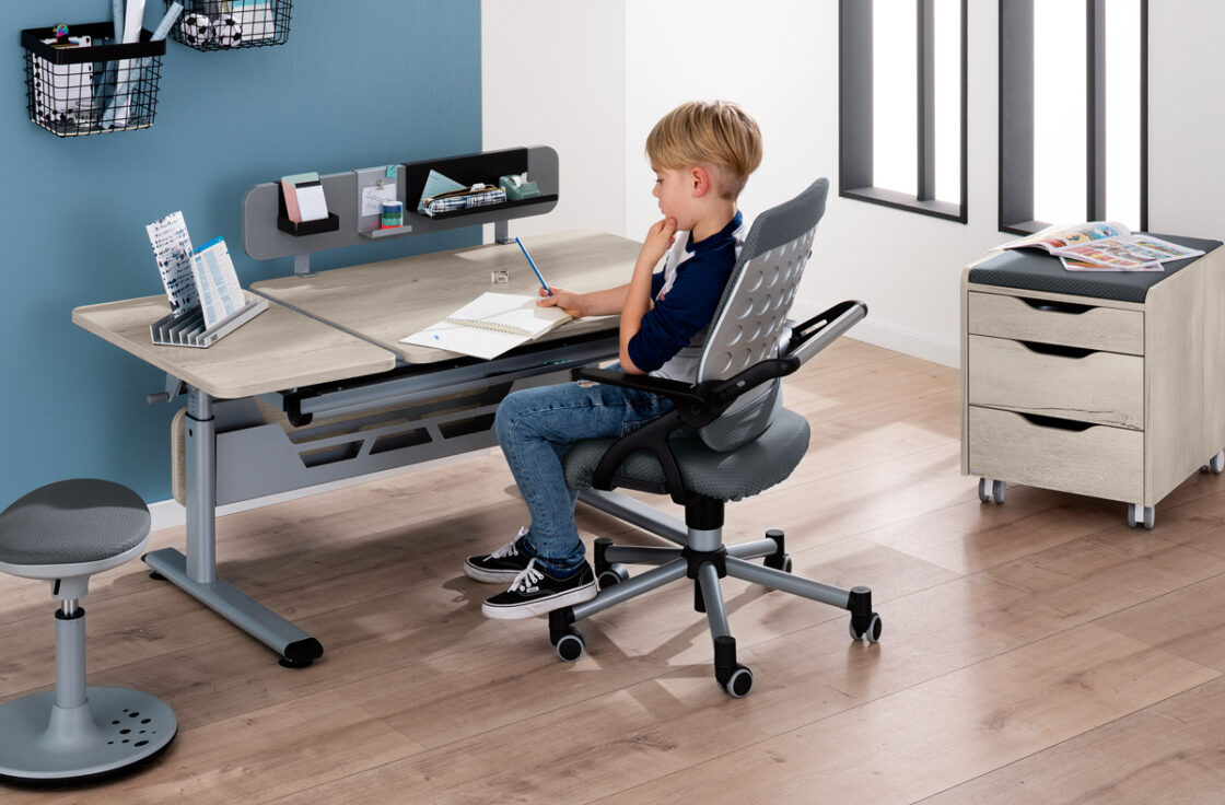 Junge sitzt am PAIDI Kinderschreibtisch Diego GT mit Funktionsboard, Rollcontainer 3S, Schreibtischstuhl Tio und Sitzhocker Speedy in einem modernen Kinderzimmer.